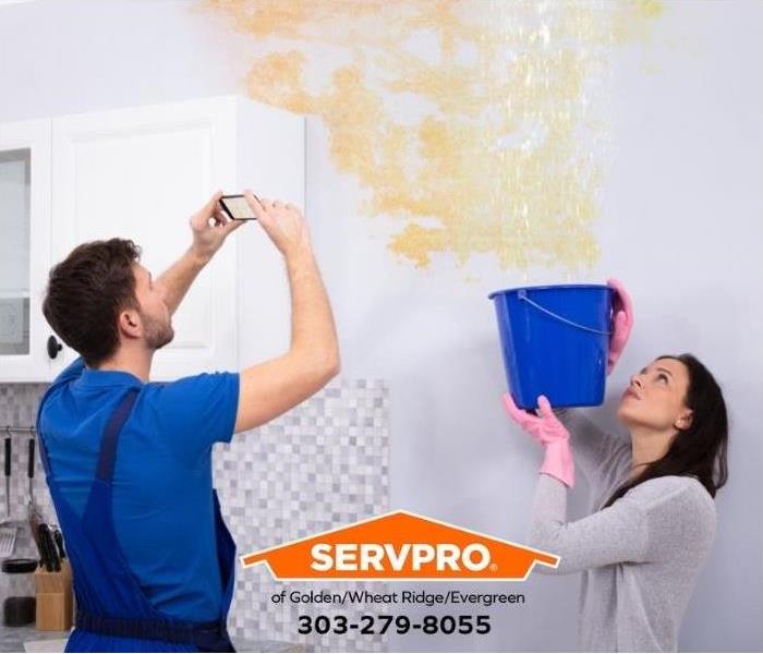 A person takes a picture of a water stain on the ceiling and wall while another person holds up a bucket to catch water drips